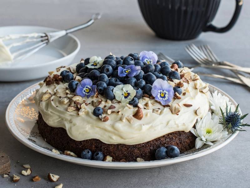 Gulrotkake med kremostfyll, toppet med blåbær, mandelflak og spiselige blomster, servert på et hvitt fat.