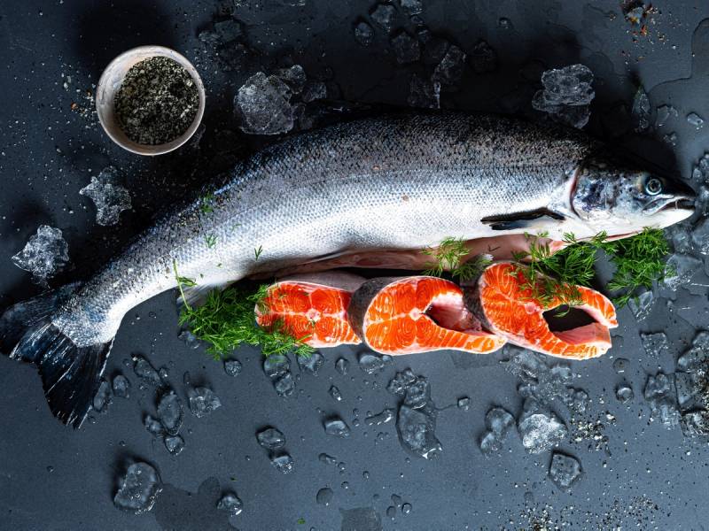 En hel fersk fisk med sitrusbiter og urter på en mørk overflate med isbiter og en liten skål med krydder eller salt i nærheten.