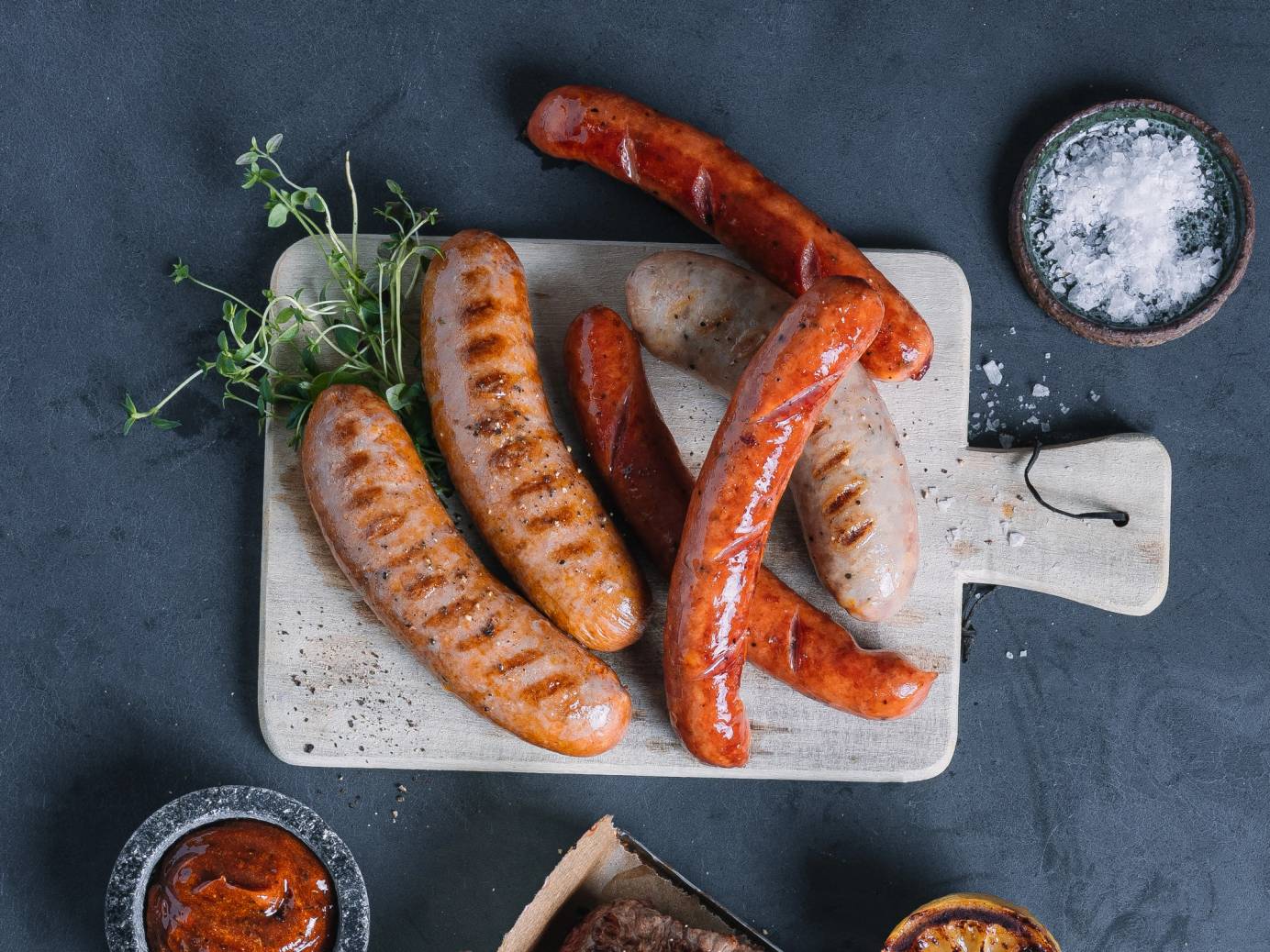 Grillpølser på et skjærebrett med noen grønne urter og små skåler med saus ved siden av.