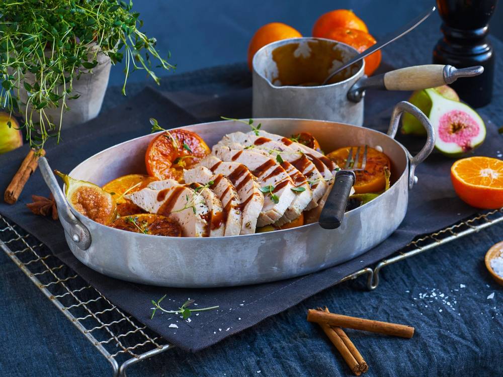 En ovnsbakt kyllingfilet med sitrus og urter på en serveringsfat, omgitt av kuttede appelsiner og en liten gryte med saus, på et mørkt bord med krydder og urter rundt.