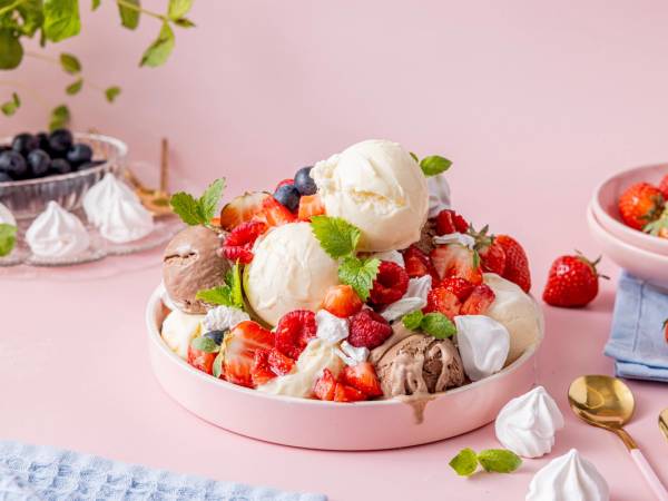En dessert med bær, marengs og iskuler i en rund, rosa serveringsskål, med flere bær og små kaker rundt på bordet.