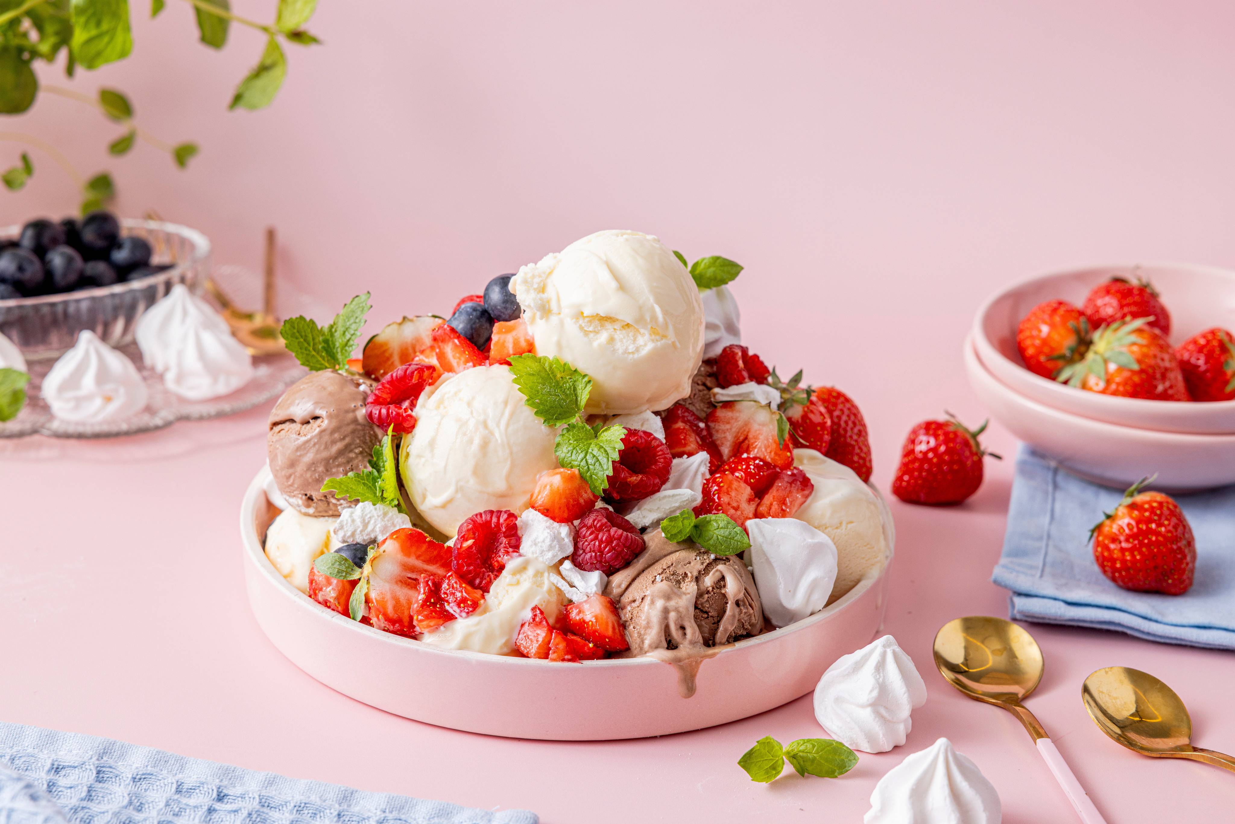 En dessert med bær, marengs og iskuler i en rund, rosa serveringsskål, med flere bær og små kaker rundt på bordet.