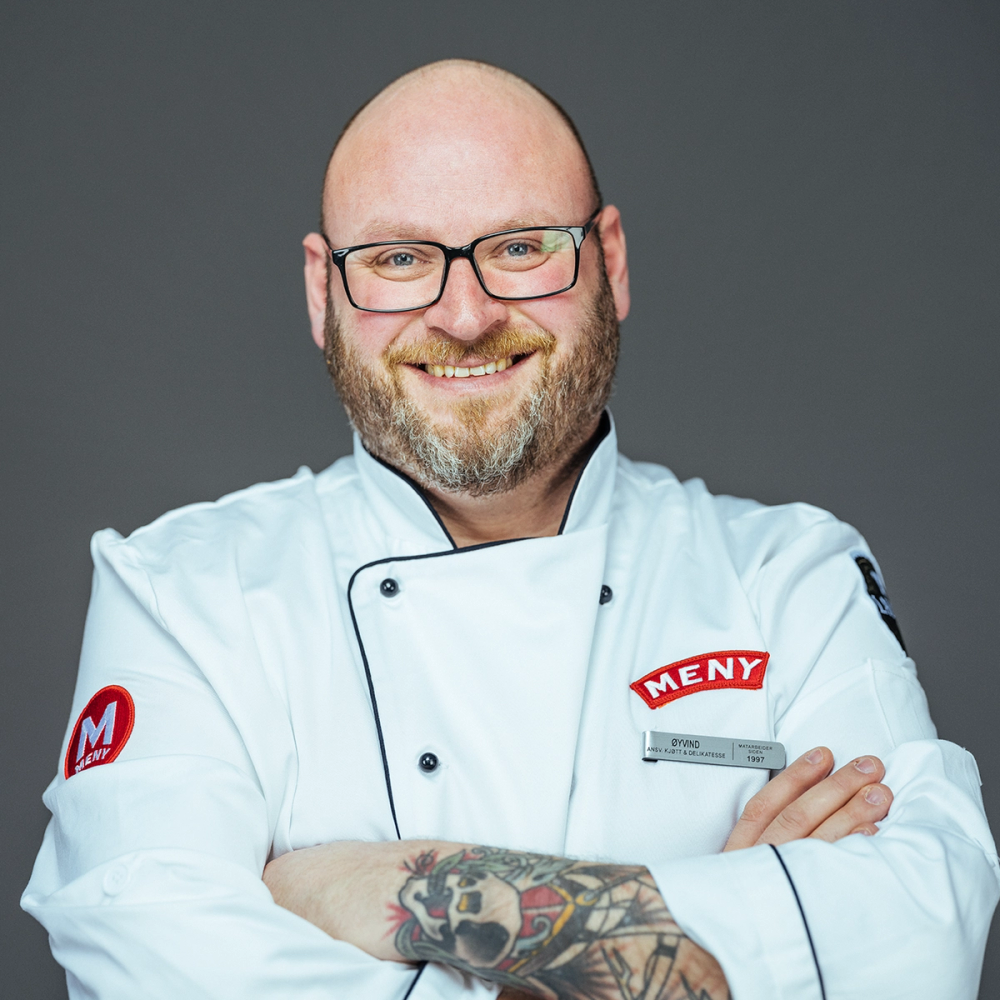 En person i hvit kokkebekledning med armer korslagt, med en tatovering på armen, står mot en grå bakgrunn.