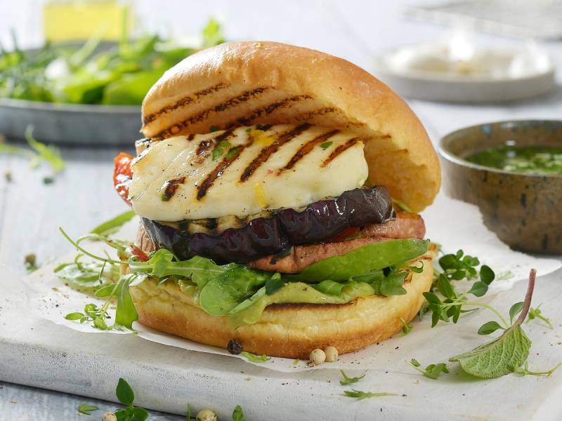 Burger med grillede halloumiost, salat og tomat i et hamburgerbrød, med en liten skål med saus ved siden av.