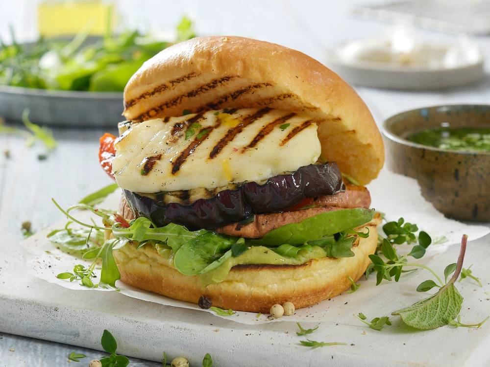 Burger med grillede halloumiost, salat og tomat i et hamburgerbrød, med en liten skål med saus ved siden av.