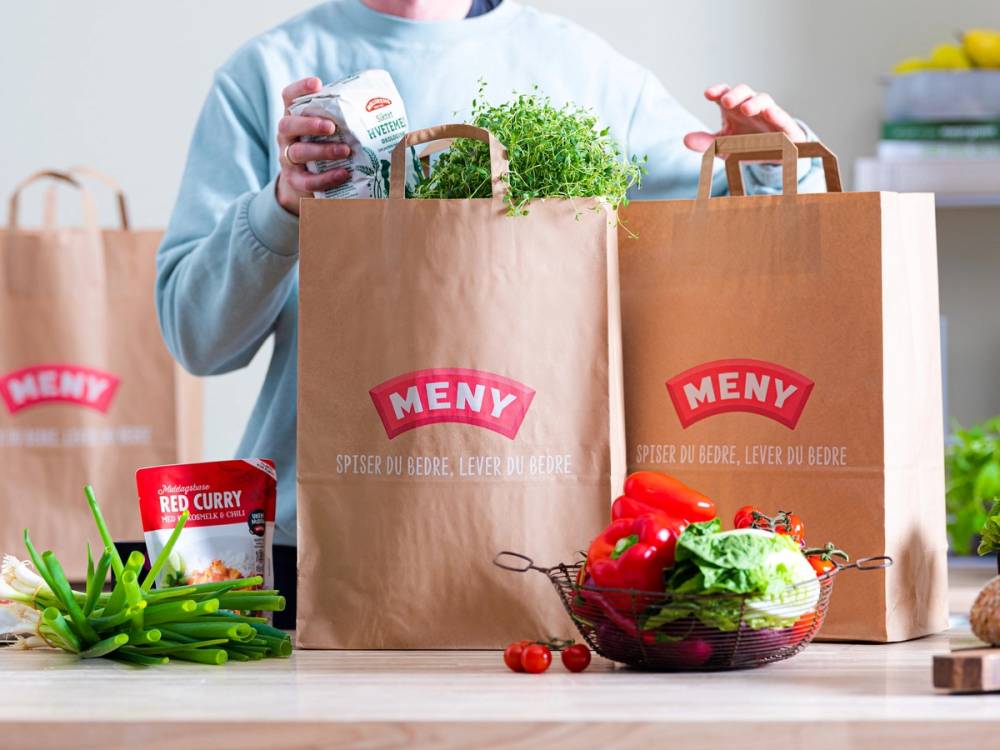 En person som pakker opp handleposer fra Meny med matvarer og grønnsaker på kjøkkenbordet.