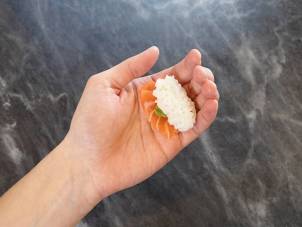 En person holder en bit nigiri-sushi med laks og en klatt med wasabi på en svart steinplate.