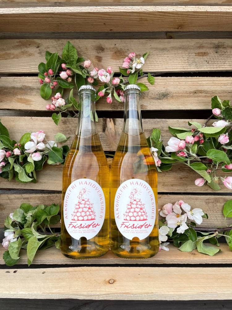 To glassflasker med eplejuice foran en blomsterdekor av rosa og hvite blomster og grønne blader på en trevegg