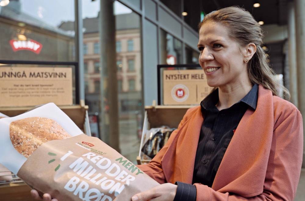 En person med bløt jakke og svart skjorte står foran en matbutikk og holder en pose med brød, mens butikkens vindu og skilt er synlige i bakgrunnen.