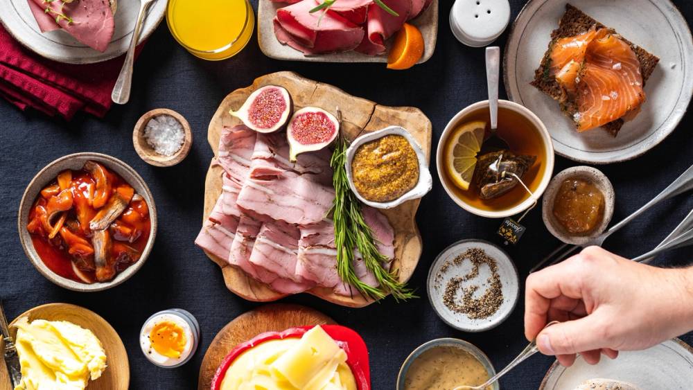 Et julefrokostbord med ulike retter som spekemat, gravlaks, og tilbehør, med en person som serverer mat.