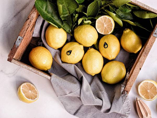 Seks sitroner med gule skinn og noen med kuttede halvdeler i en trekasse med grønne blader rundt.