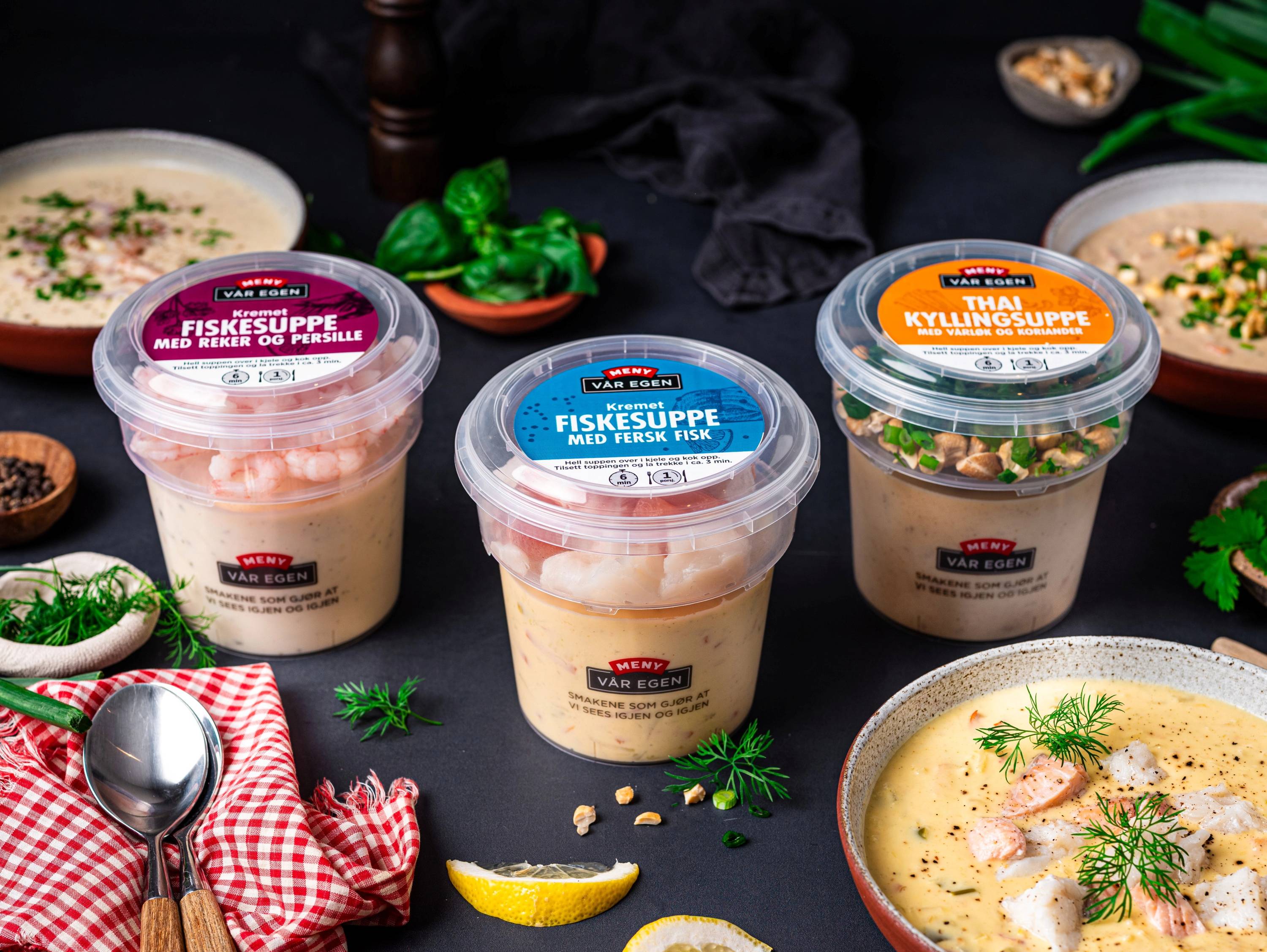 To kopper med fiskesuppe og en skål med kyllingsuppe omgitt av grønnsaker og sitronbiter på et bord.