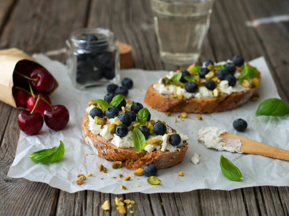 Ricotta med blåbær, pistasj og ferske kirsebær på et stykke brød, med et glass vann og en liten krukke med bær i bakgrunnen.