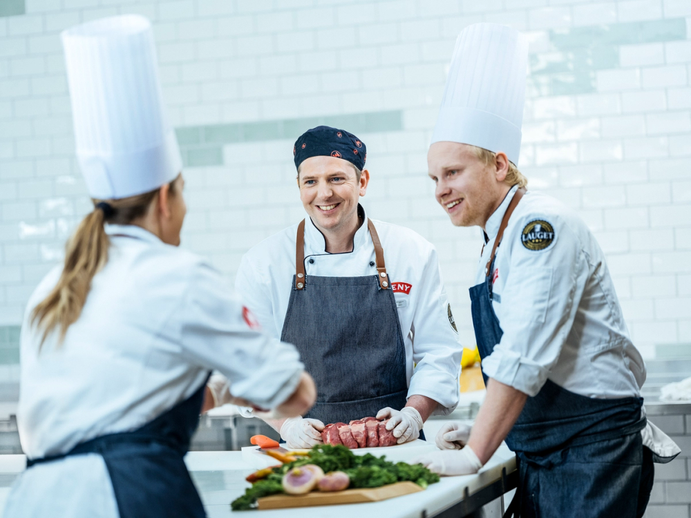 Tre kokker i profesjonelle uniformer og kokkehatter som jobber sammen på kjøkkenet med kjøtt og grønnsaker foran seg.