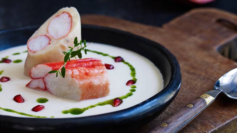 Kongekrabbe med jordskokksuppe servert i en svart bolle med pynt av urter og dråper av saus rundt.