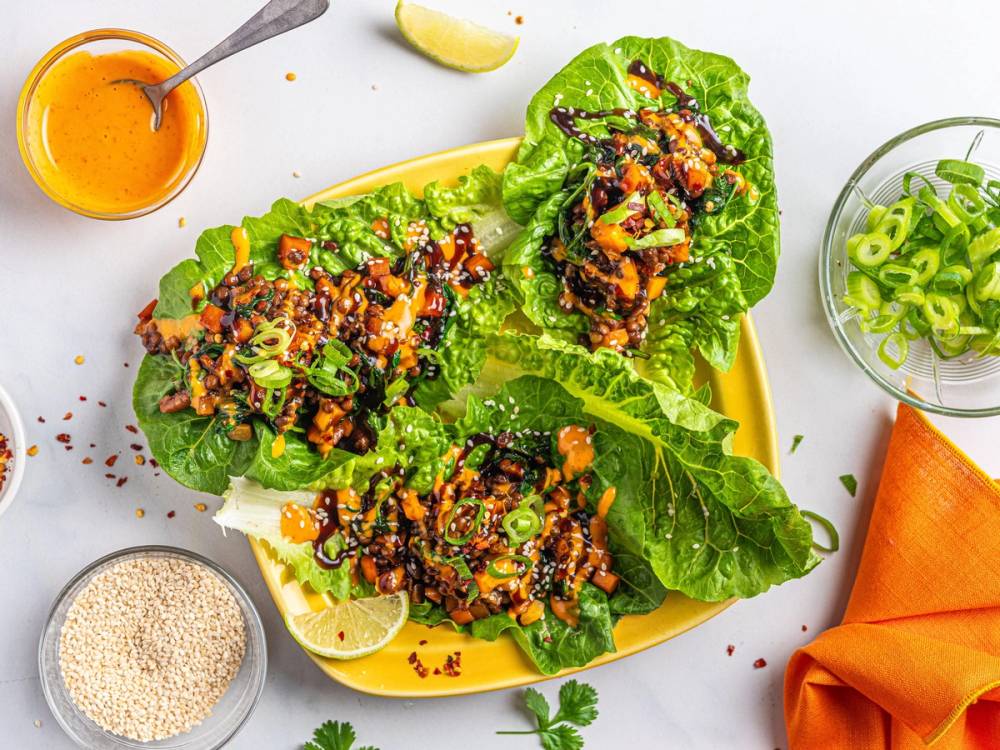 Asiatiske tacos med kjøtt, grønnsaker og grønne løk på en gul tallerken, omgitt av en liten bolle med saus, en kopp med sesamfrø, og en liten skål med grønne løk, på et hvitt bord.