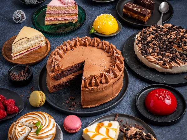 Et utvalg av kaker og desserter på en butikkdisk, inkludert sjokoladekake, ostekake, fruktterter og småkaker.