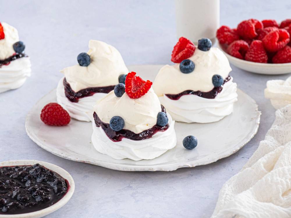 Små pavlovaer med krem, bær og rips på et hvitt fat, med en skål med bær i bakgrunnen.