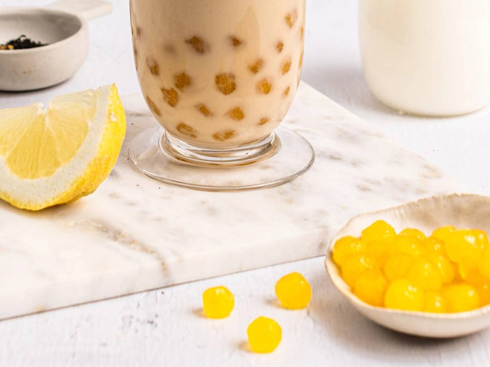 Et glass med te og melk, en skål med gule små kuler, og en sitronskive på en hvit overflate.