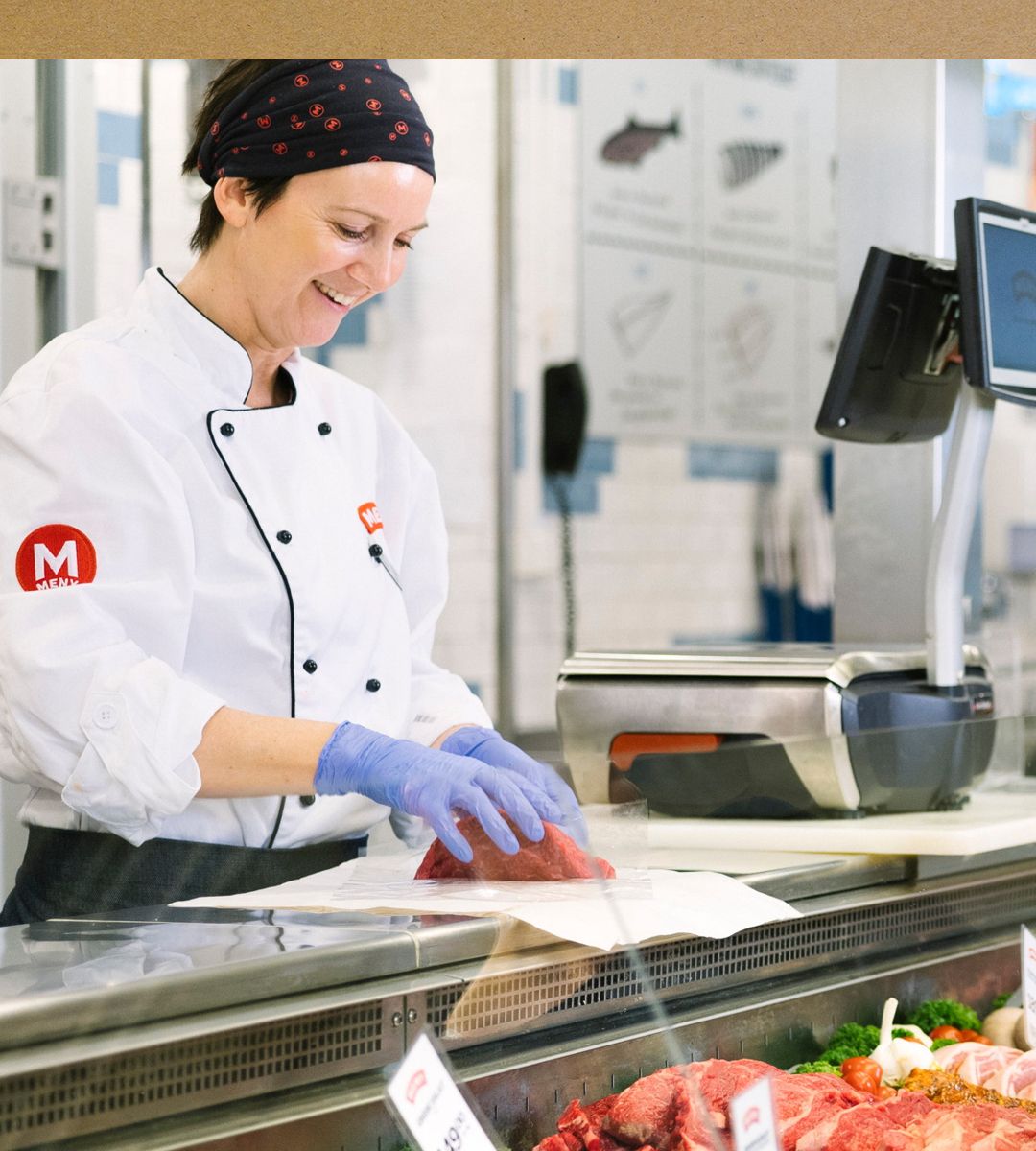 En kokk i hvit uniform og blå hansker skjærer kjøtt i en butikk eller på et marked, med kjøtt og utstyr rundt seg.