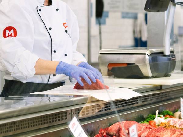 En kokk i hvit uniform og blå hansker skjærer kjøtt i en butikk eller på et marked, med kjøtt og utstyr rundt seg.