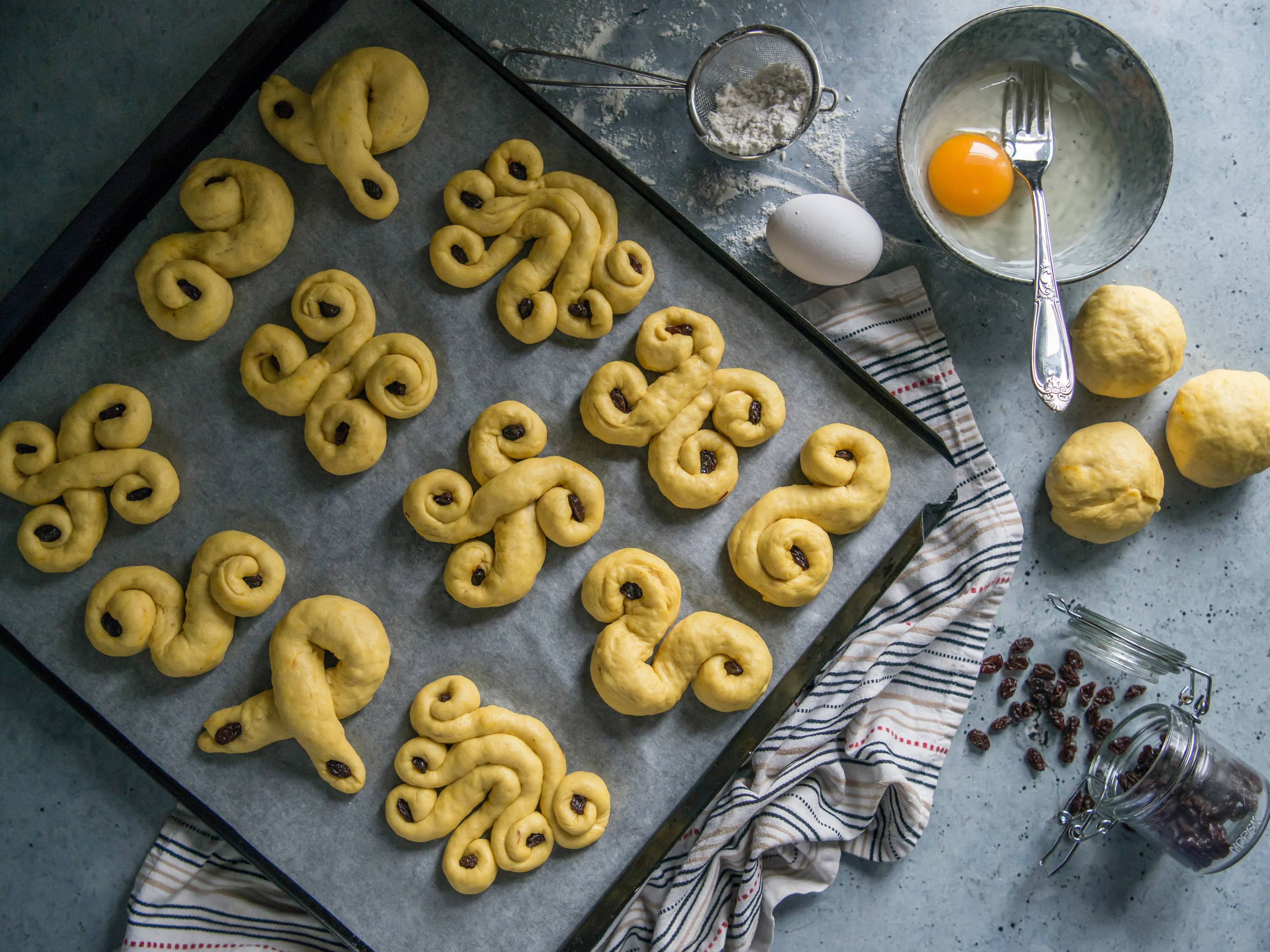 Ulike lussekatter på en bakeplate, klare for steking, med ingredienser og kjøkkenutstyr rundt.