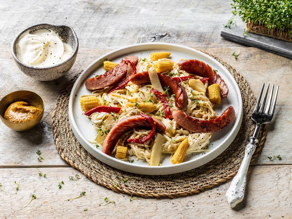 Wok med grønnsaker, nudler og kjøttpølse på en hvit tallerken, servert med en skje og et glass med en hvit drikk, på et trebenke-aktig underlag.