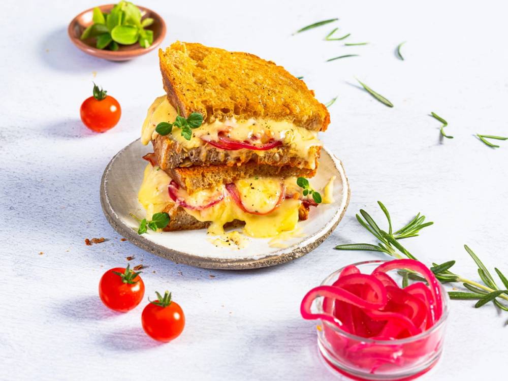 En toast med ost og skinke på en tallerken, omgitt av cherrytomater og grønne urter.