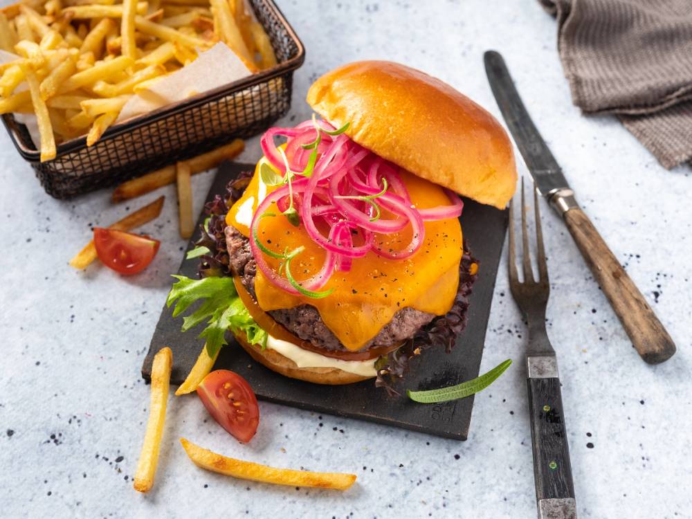 Cheddarost på en burger med salat, løk og tomater, servert på en svart tallerken med pommes frites ved siden av.