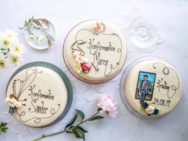 Tre konfirmasjonskaker med dekorasjoner og tekst, plassert på et hvitt bord med blomster og servietter.