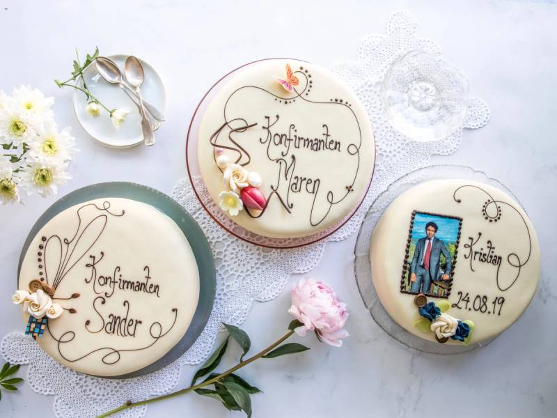 Tre konfirmasjonskaker med dekorasjoner og tekst, plassert på et hvitt bord med blomster og servietter.