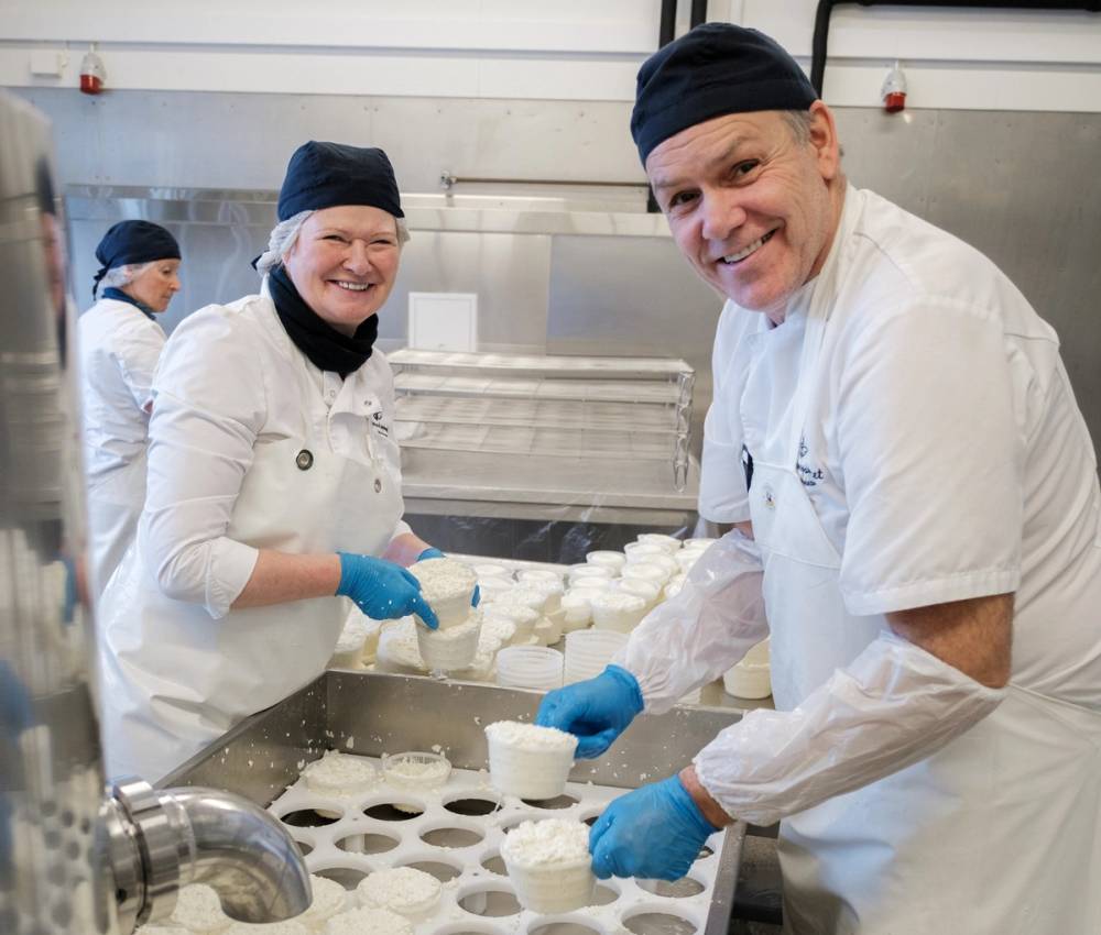 To personer i hvite kokkerklær jobber med ost i et kjøkken, med en person som bruker blå hansker, og en annen som har på seg en svart lue.