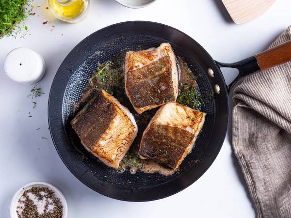 Tre skreifileter stekes i en panne med litt smør, omgitt av krydder og olivenolje på et kjøkkenbord.