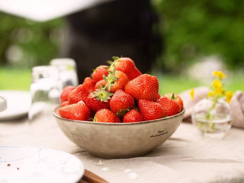 En skål med ferske jordbær på et utendørs bord, med grønne trær i bakgrunnen.