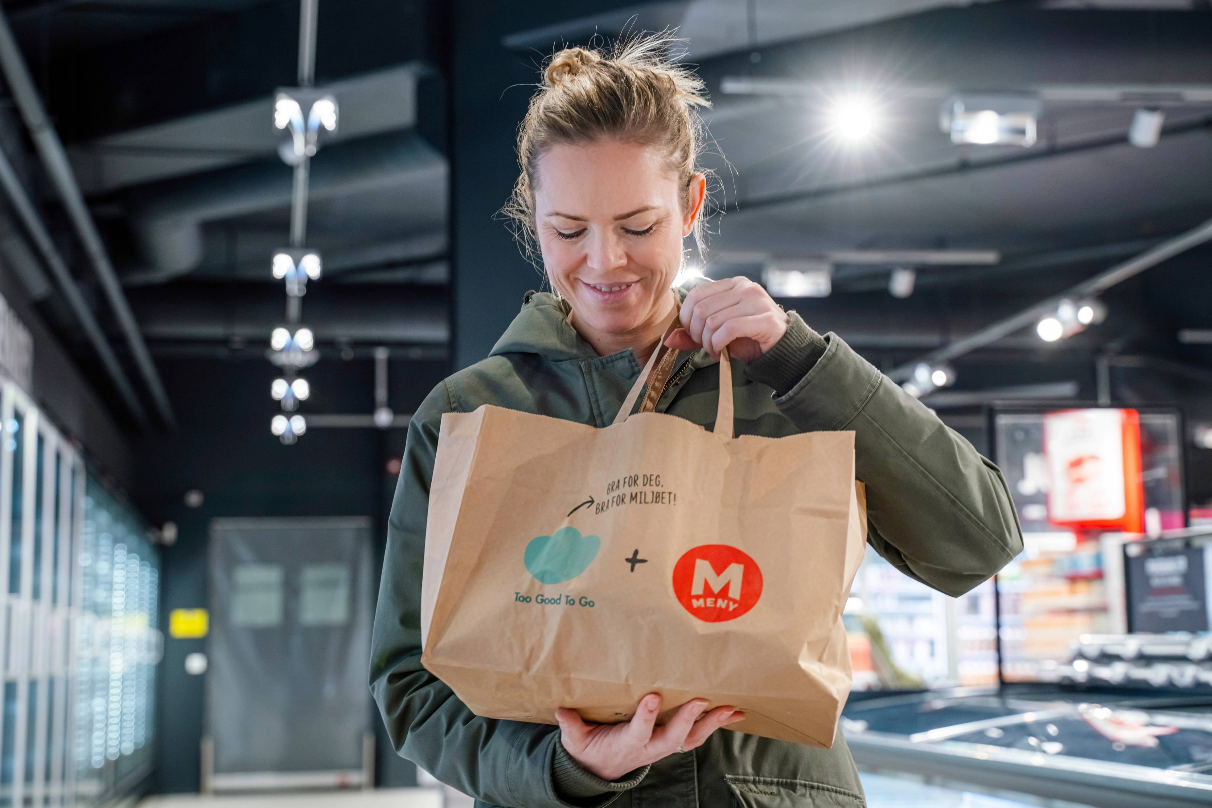 En person i en butikk holder en papirpose med en logo og et pluss-tegn, med hyller og lys i bakgrunnen.