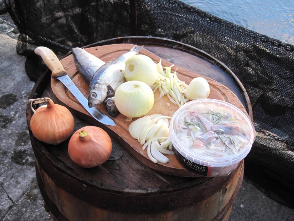 Løk, en hel fisk, og en plastboks med fisk og grønnsaker på et skjærebrett, plassert på en trebord utendørs.