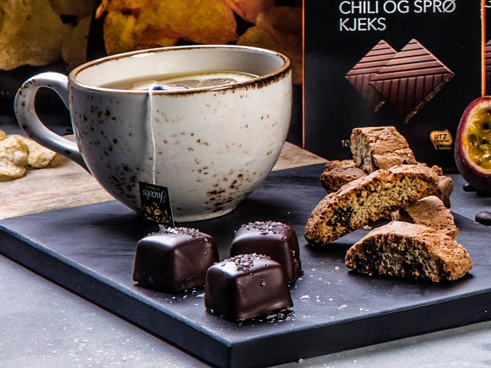 En kopp med kaffe og et utvalg sjokolade og kjeks på et serveringsfat, med sjokoladeplater i bakgrunnen.