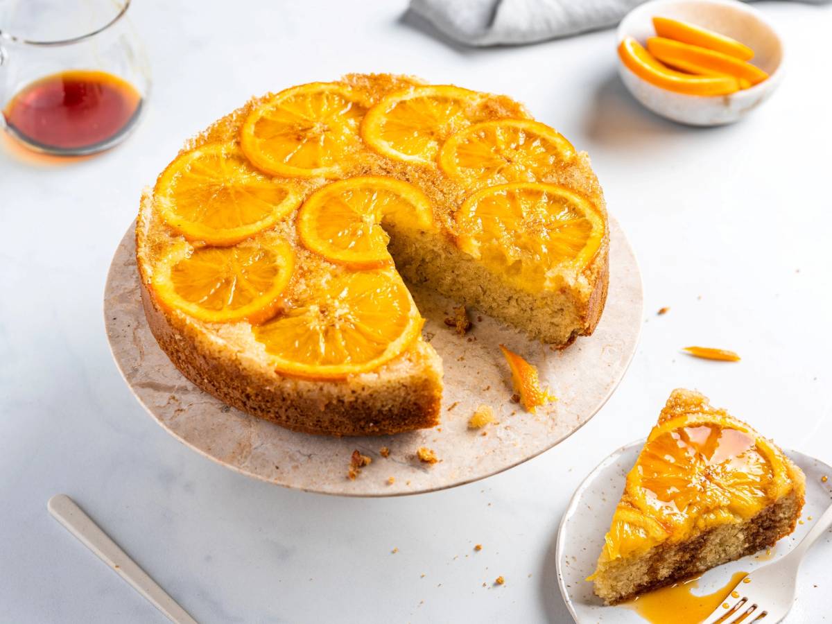 En appelsinkake med skiver av appelsin på toppen, opp ned, med en skive tatt ut og plassert på en liten tallerken ved siden av.