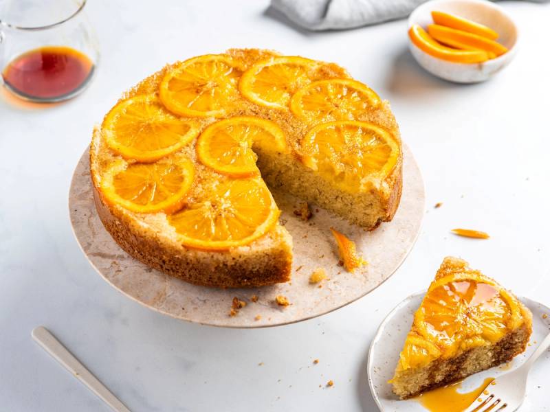 En appelsinkake med skiver av appelsin på toppen, opp ned, med en skive tatt ut og plassert på en liten tallerken ved siden av.