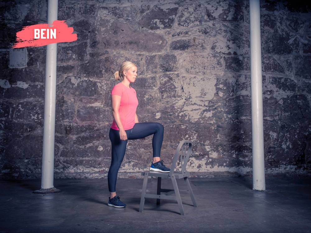 En kvinne står med en fot på en stol og holder balansen i et treningsrom med en grå vegg i bakgrunnen.