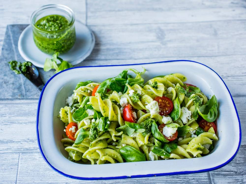 Pastasalat med grønnsaker og fetaost i en hvit serveringsskål med en glass med grønn dressing ved siden av, på et lyst bord.