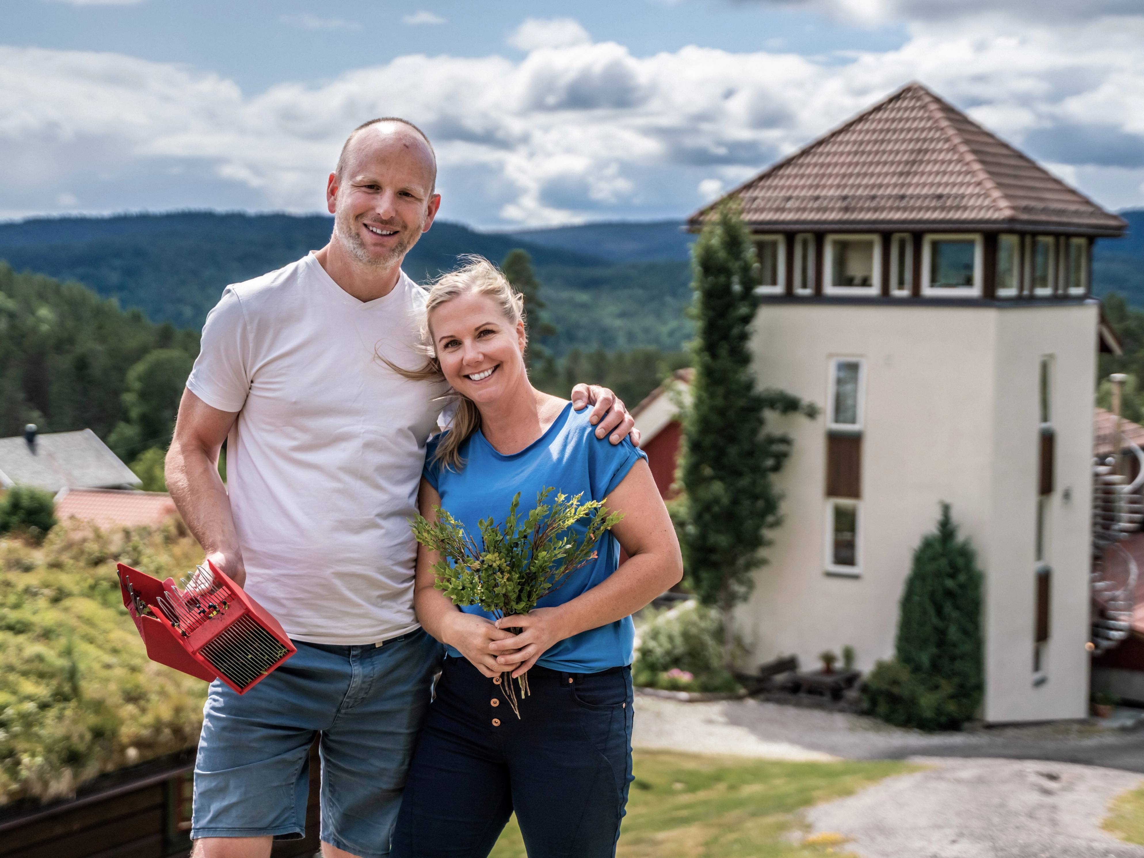 To personer står sammen foran et hus med utsikt over landskap og fjell i bakgrunnen, en av dem holder en bok eller mappe.