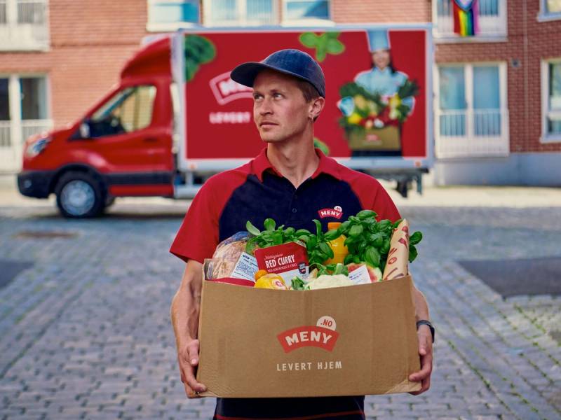 En person i rød uniform holder en stor eske med matvarer foran en rød leveringsbil, med boligblokker i bakgrunnen.