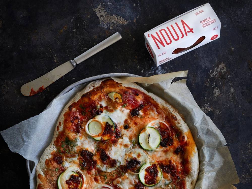 En pizza med nduja, skiver av zucchini og tomatsaus, på en stekeplate med en kniv og en pakke med nduja ved siden av.