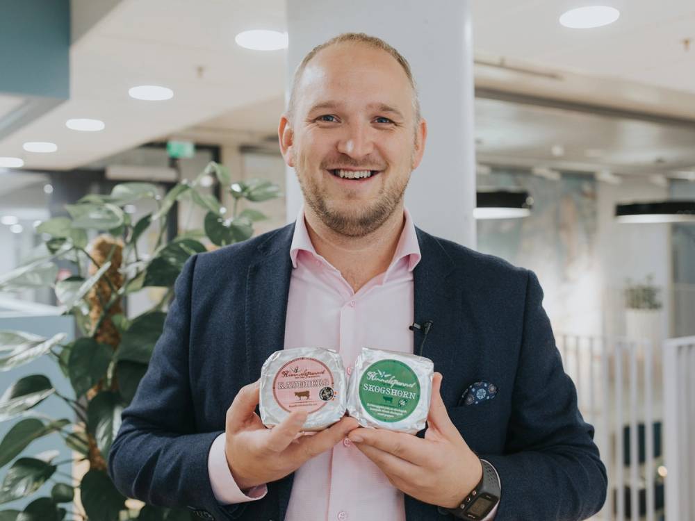 Person holder to produkter, med en bakgrunn som antyder en utstilling eller premieutdeling, trolig relatert til Norges største lokalmatpris.