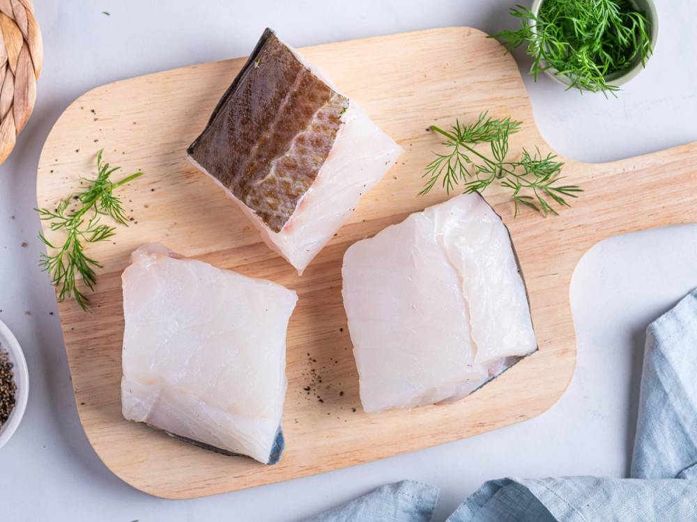 Tre stykker skreifisk ligger på et rundt trebrett med noen grønne urter ved siden av.