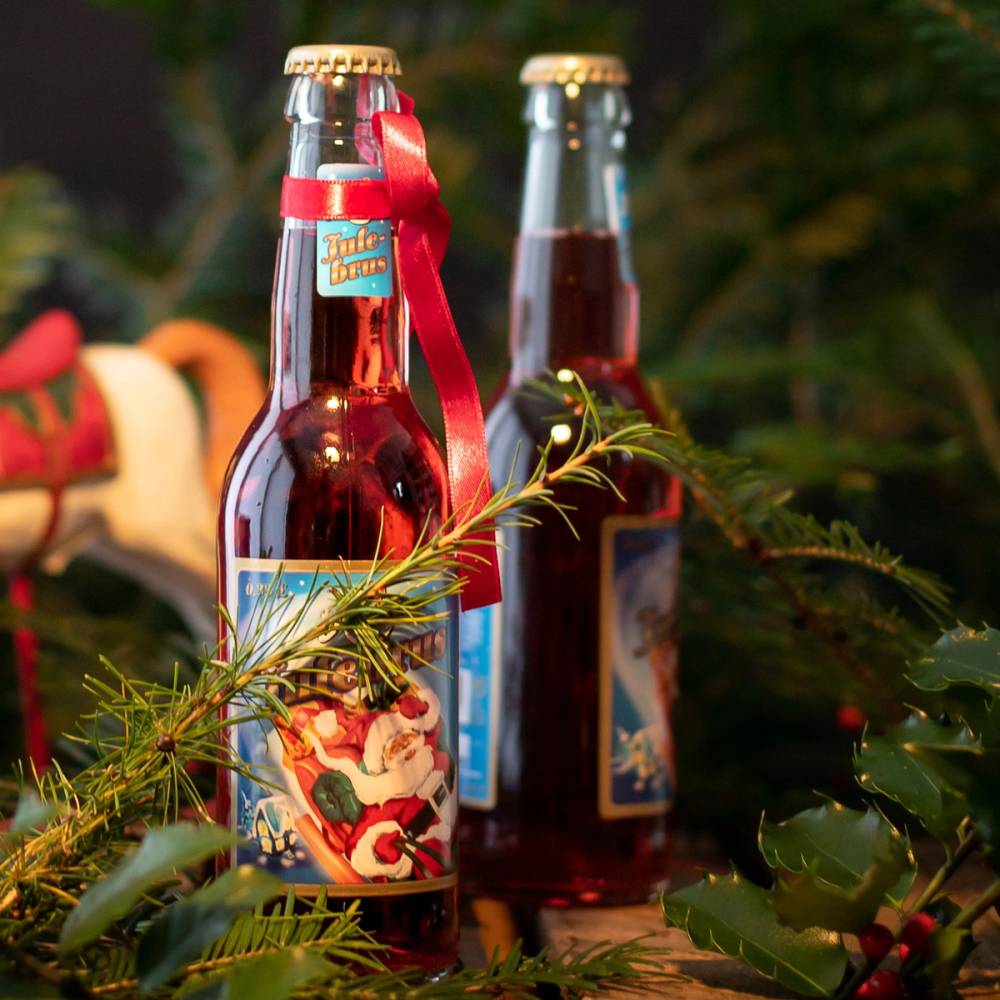 To flasker med Hansa julebrus plassert blant julegrener og pynt, med en julefigur i bakgrunnen.