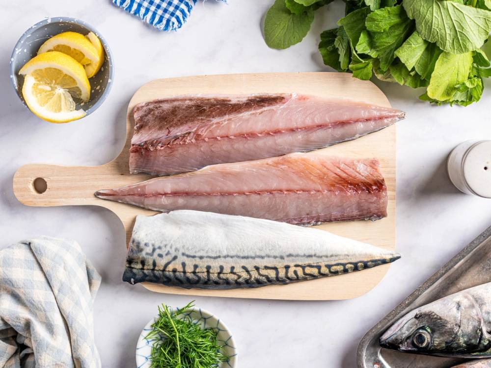 Ulike typer av makrellfileter og fersk fisk på et kjøkkenbord med grønnsaker og sitron, klar til tilberedning.