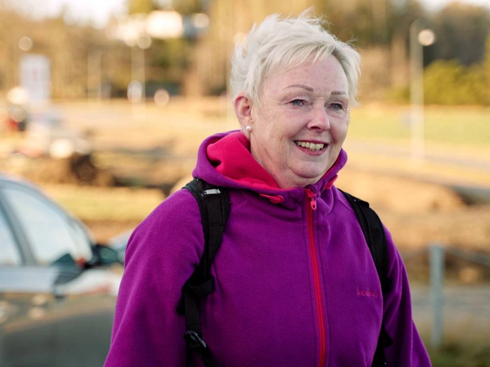 En person med lyst hår og lilla jakke står utendørs med en ryggsekk, med en park eller gate i bakgrunnen.