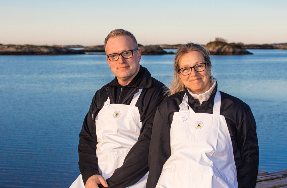 To personer står på en brygge ved vannet, begge iført hvite forkle og svarte jakker, med vann og en klar himmel i bakgrunnen.